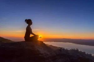 Woman practicing mindful meditation on a mountain at sunset, promoting wellness and mental health.